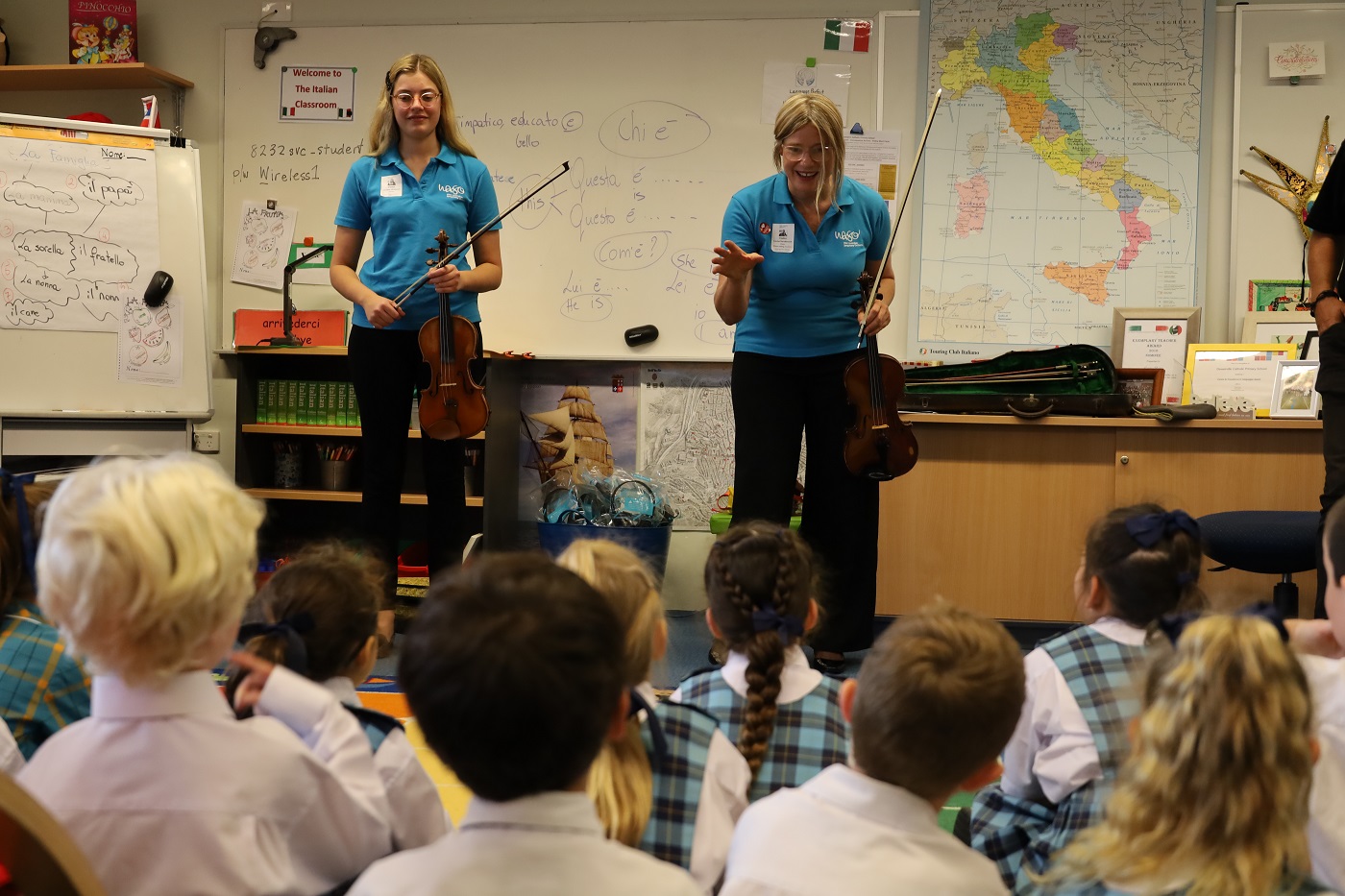A violinist and violist giving a presentation in a primary school classroom, while students seated on the floor watch.