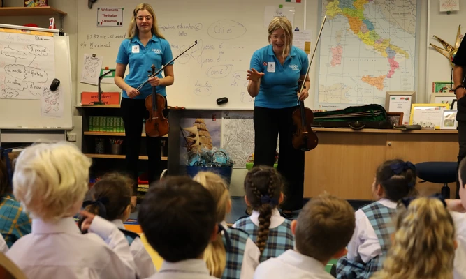 A violinist and violist giving a presentation in a primary school classroom, while students seated on the floor watch.