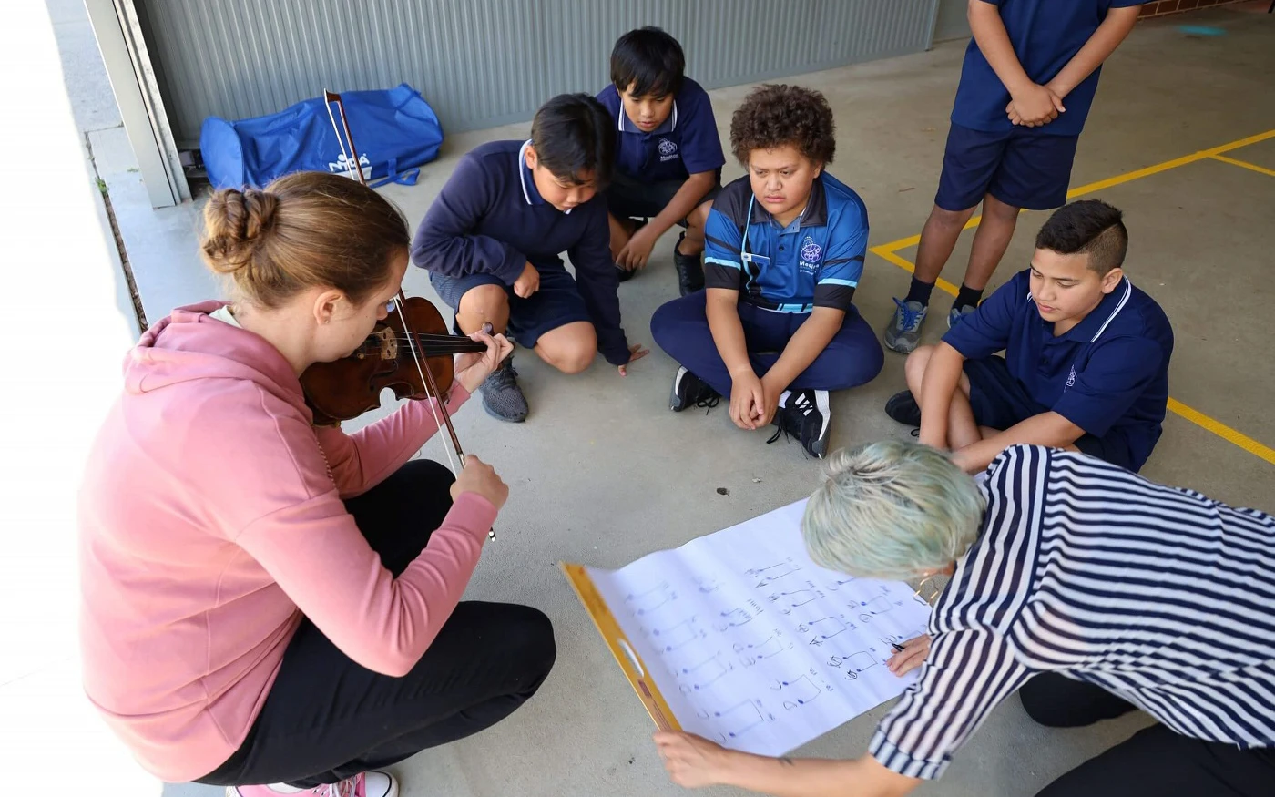 A violinist and teacher sit on the ground with a group of primary school students, reading a large page of sheet music together.