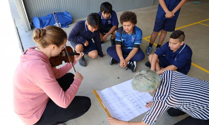 A violinist and teacher sit on the ground with a group of primary school students, reading a large page of sheet music together.