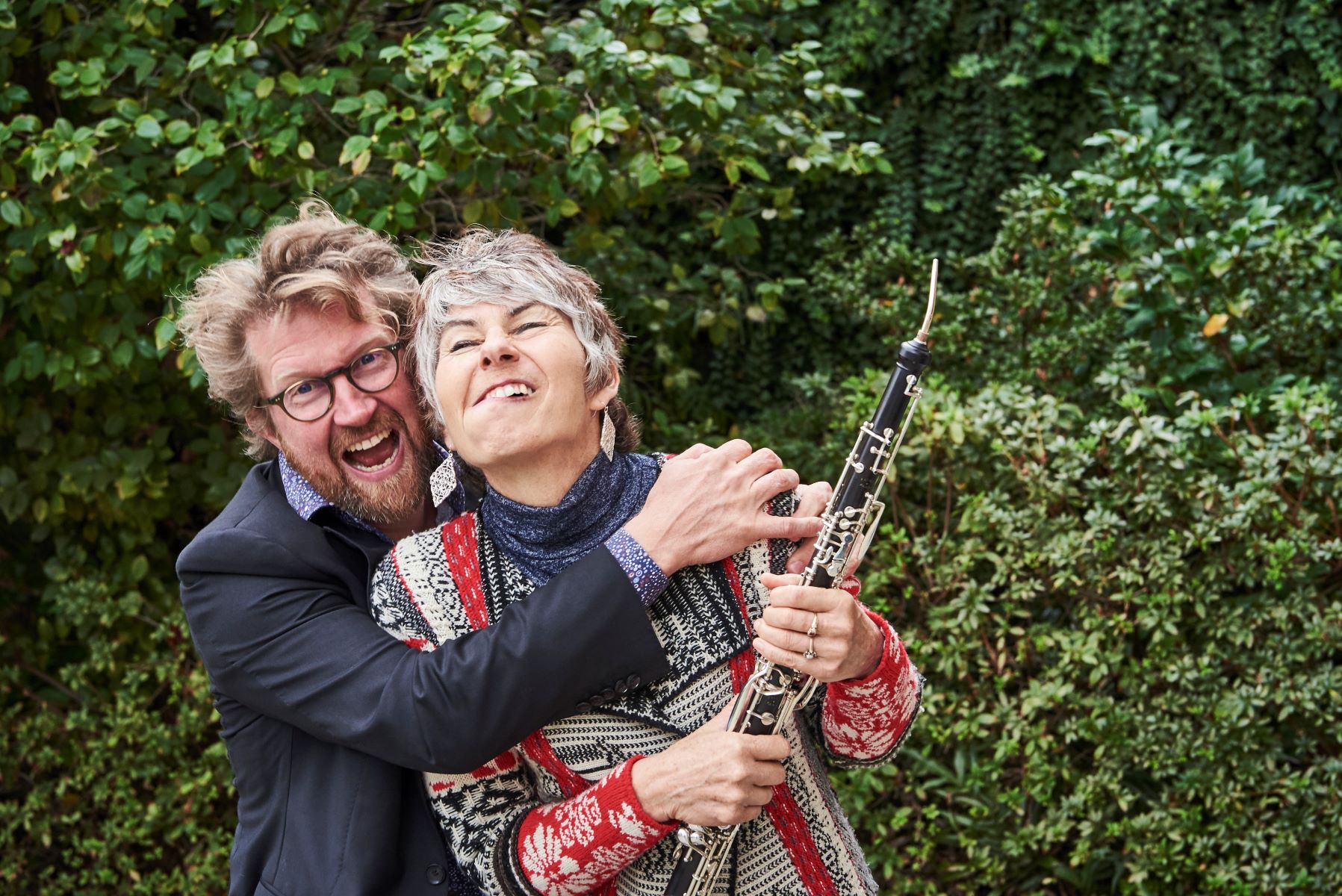 Leanne Glover and Iain Grandage embrace and pull faces for a silly photo, surrounded by leafy garden greenery. Leanne is holding an oboe.