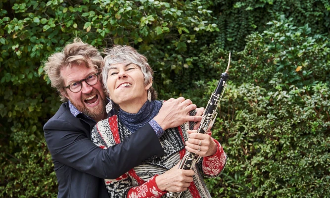Leanne Glover and Iain Grandage embrace and pull faces for a silly photo, surrounded by leafy garden greenery. Leanne is holding an oboe.
