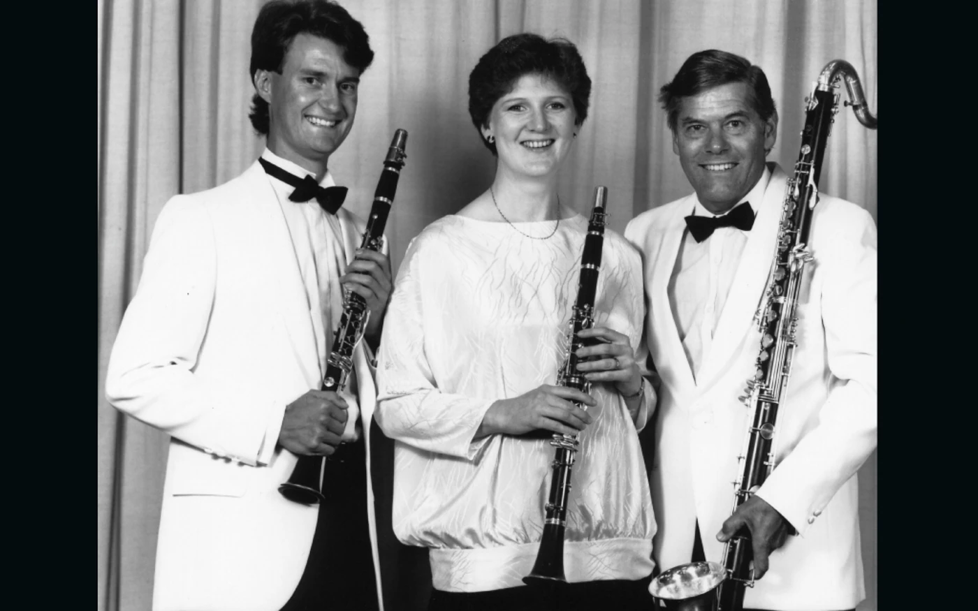 Three woodwind musicians pose with clarinets and bass clarinet in a formal, black & white group portrait.