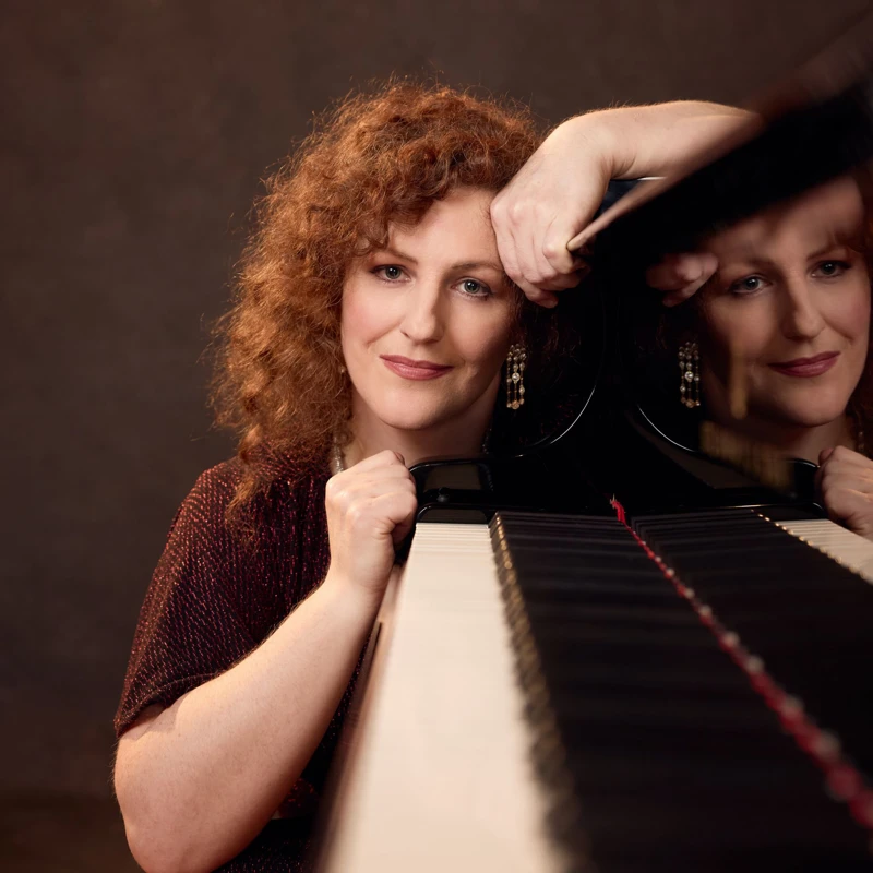 Headshot of Tamara-Anna Cislowska wearing a sparkly dark red dress, posing beside a grand piano.