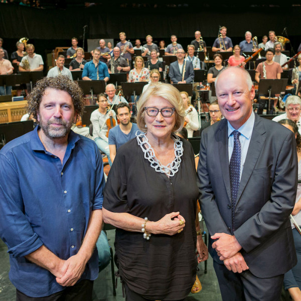 Asher Fisch, Janet Holmes à Court, and Richard Goyder smile at the camera with the orchestra in the background.