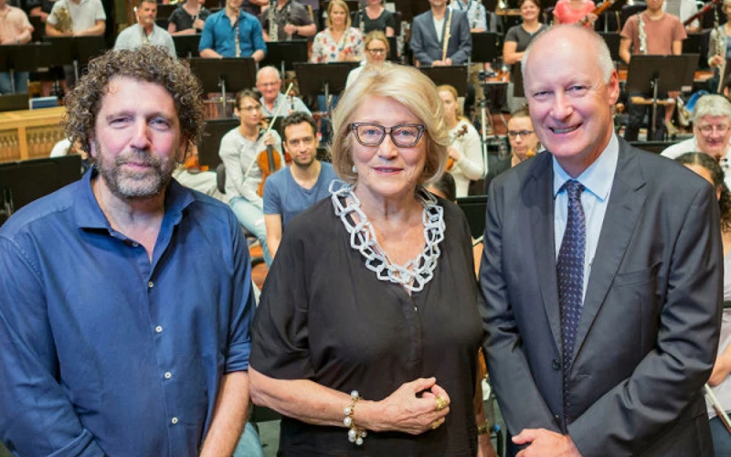 Asher Fisch, Janet Holmes à Court, and Richard Goyder smile at the camera with the orchestra in the background.