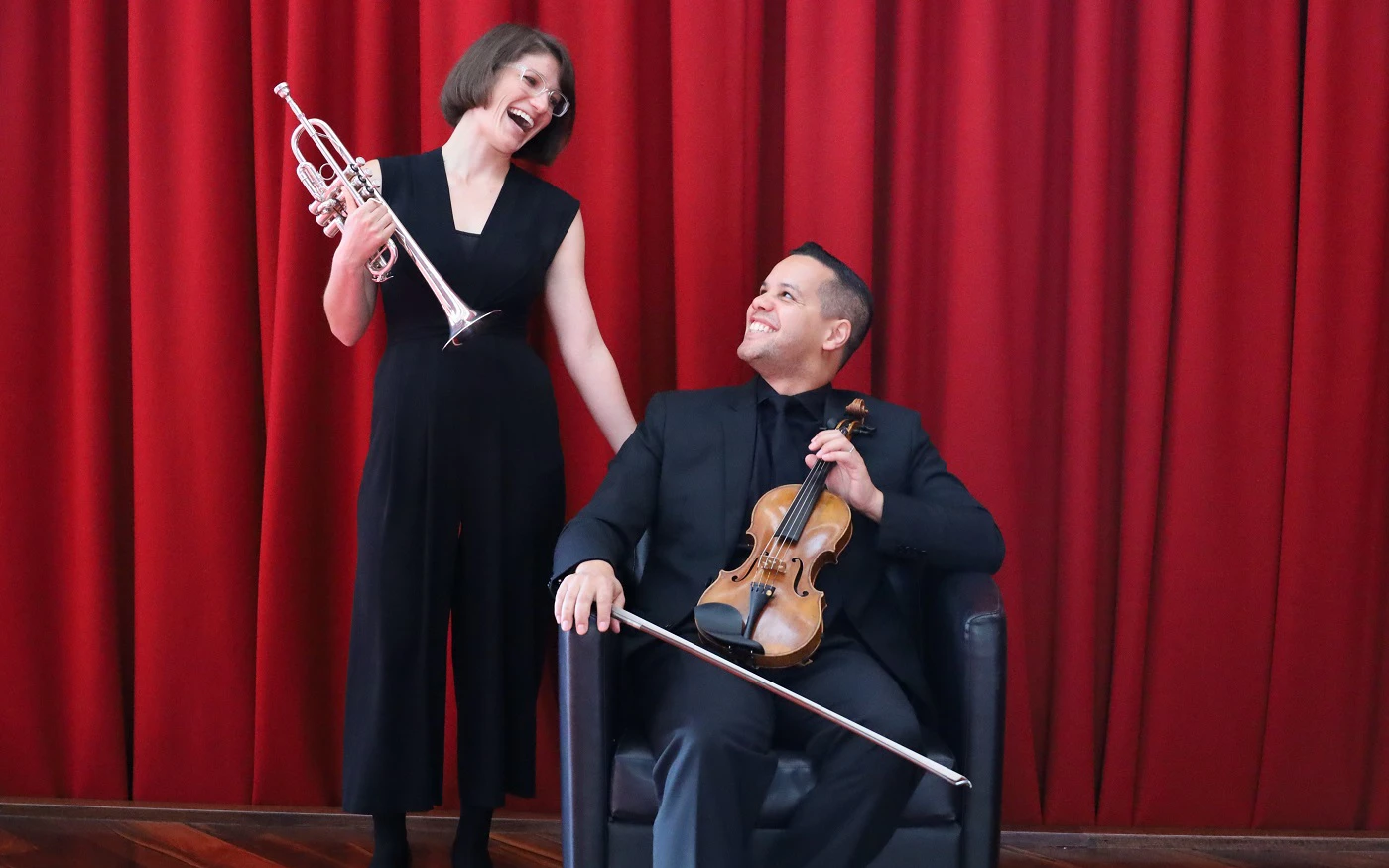 Jenna Smith (trumpet) and Graeme Norris (violin) smiling at each other in front of a red curtain backdrop, holding their instruments.