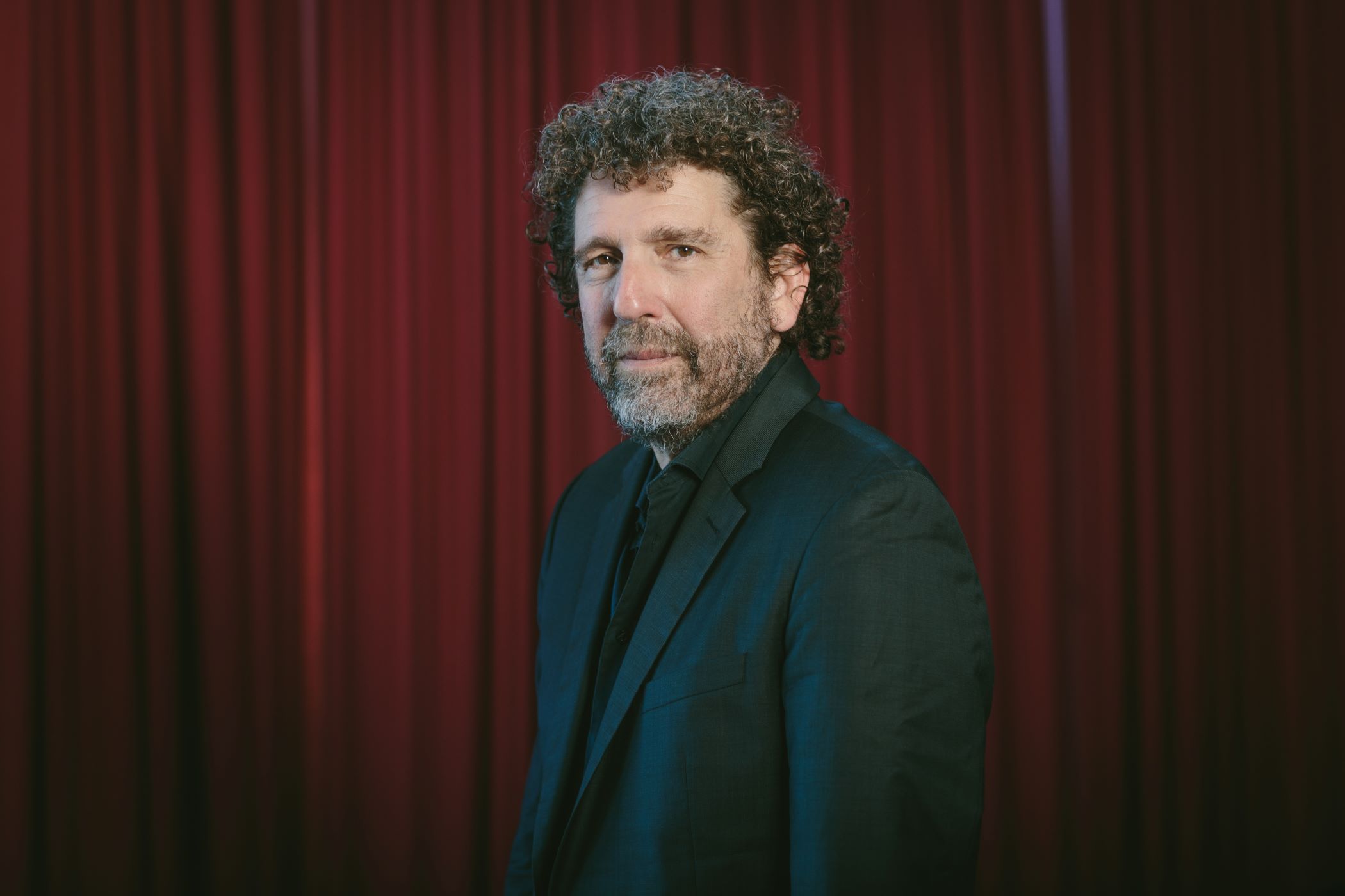 Headshot of Asher Fisch wearing a black suit and tie, against a red curtain backdrop.