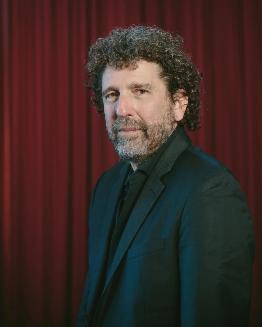 Headshot of Asher Fisch wearing a black suit and tie, against a red curtain backdrop.