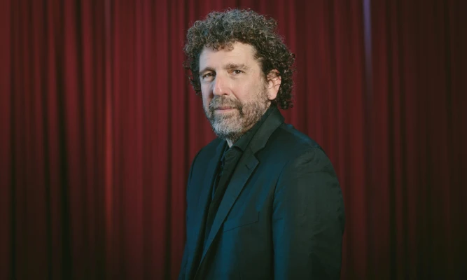 Headshot of Asher Fisch wearing a black suit and tie, against a red curtain backdrop.