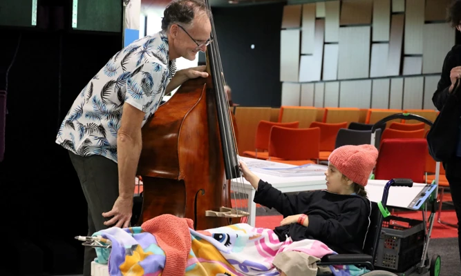 A double bass player showing his instrument to a young hospital patient in a wheelchair.