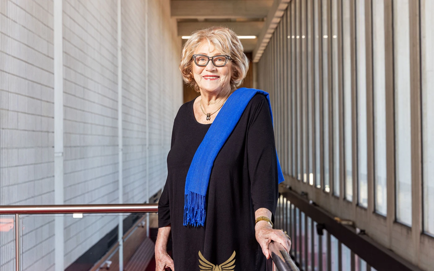 Janet Holmes à Court wearing a black dress, blue scarf and glasses, standing on a mezzanine at Perth Concert Hall.