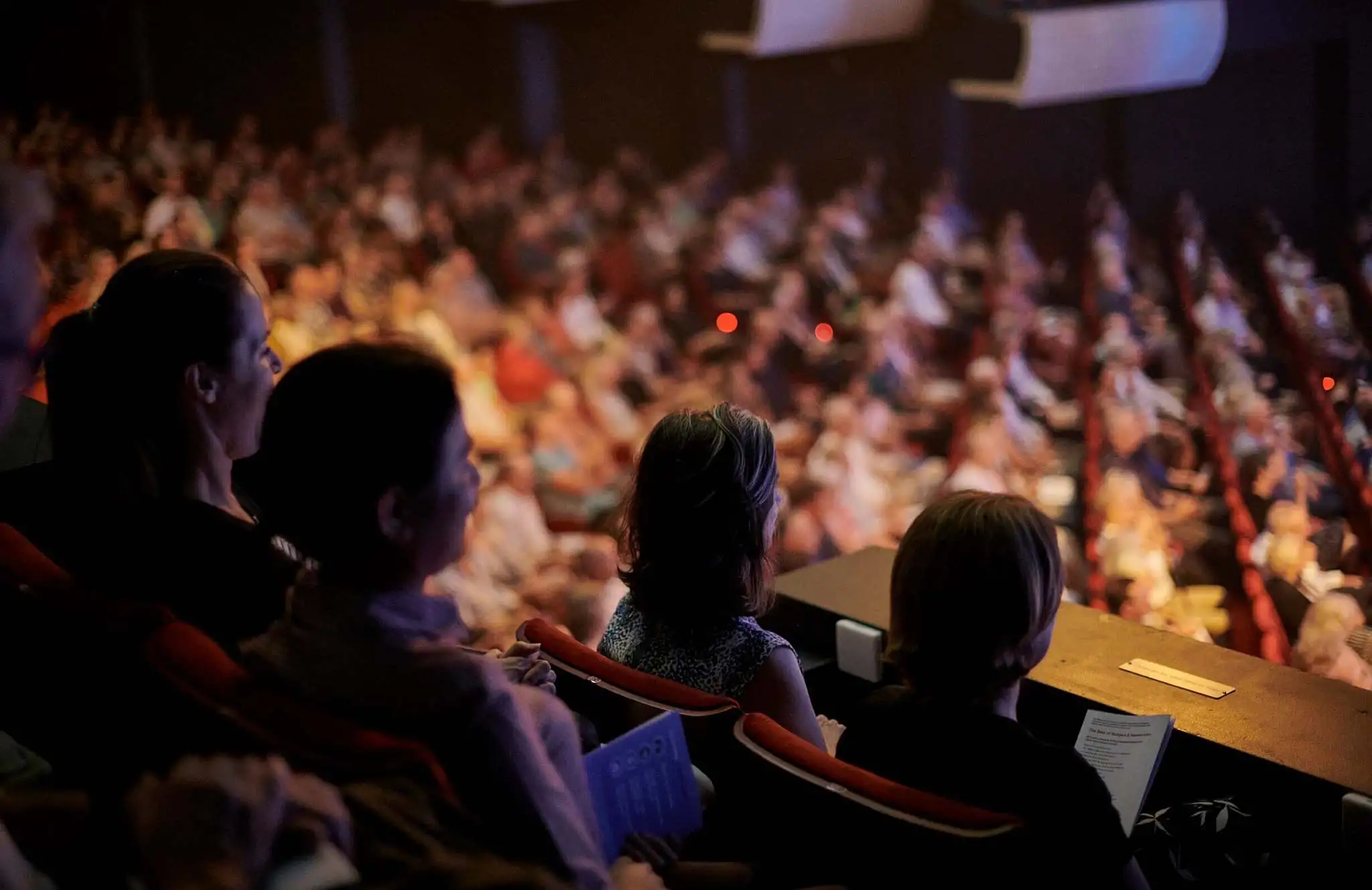 Audience members seated in the balcony of Perth Concert Hall watch a live orchestral performance, with the brightly lit stage out of frame and the rest of the auditorium softly blurred in the background.