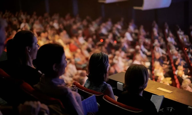 Audience members seated in the balcony of Perth Concert Hall watch a live orchestral performance, with the brightly lit stage out of frame and the rest of the auditorium softly blurred in the background.