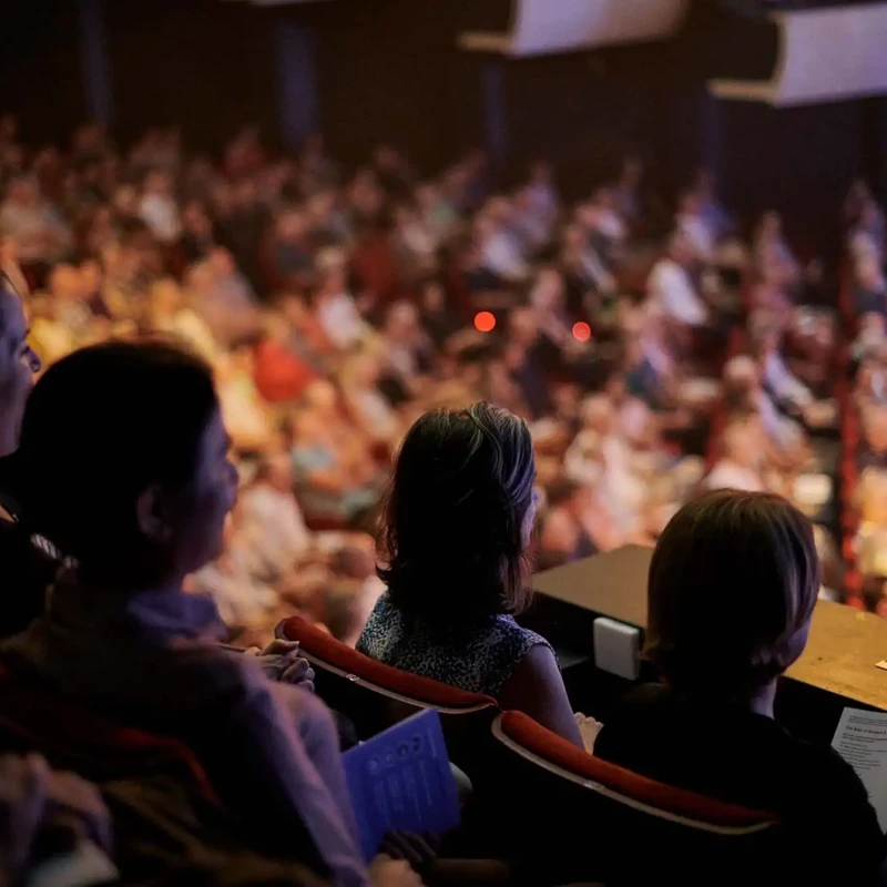 Audience members seated in the balcony of Perth Concert Hall watch a live orchestral performance, with the brightly lit stage out of frame and the rest of the auditorium softly blurred in the background.