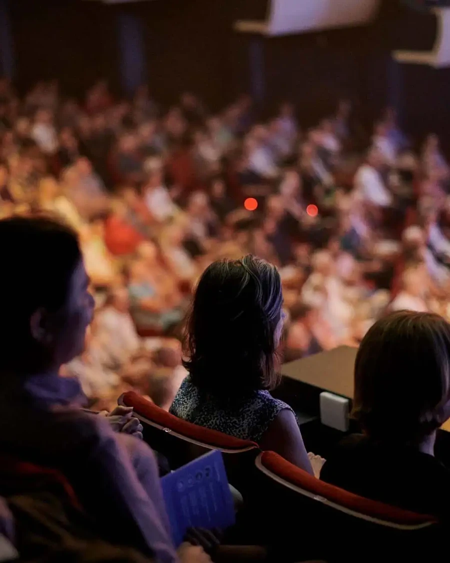 Audience members seated in the balcony of Perth Concert Hall watch a live orchestral performance, with the brightly lit stage out of frame and the rest of the auditorium softly blurred in the background.