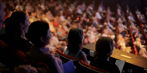 Audience members seated in the balcony of Perth Concert Hall watch a live orchestral performance, with the brightly lit stage out of frame and the rest of the auditorium softly blurred in the background.
