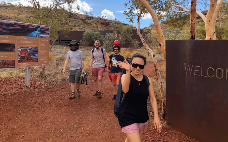 Four people dressed in hiking gear walking along a red dirt path near a 'Welcome' sign, smiling at the camera.