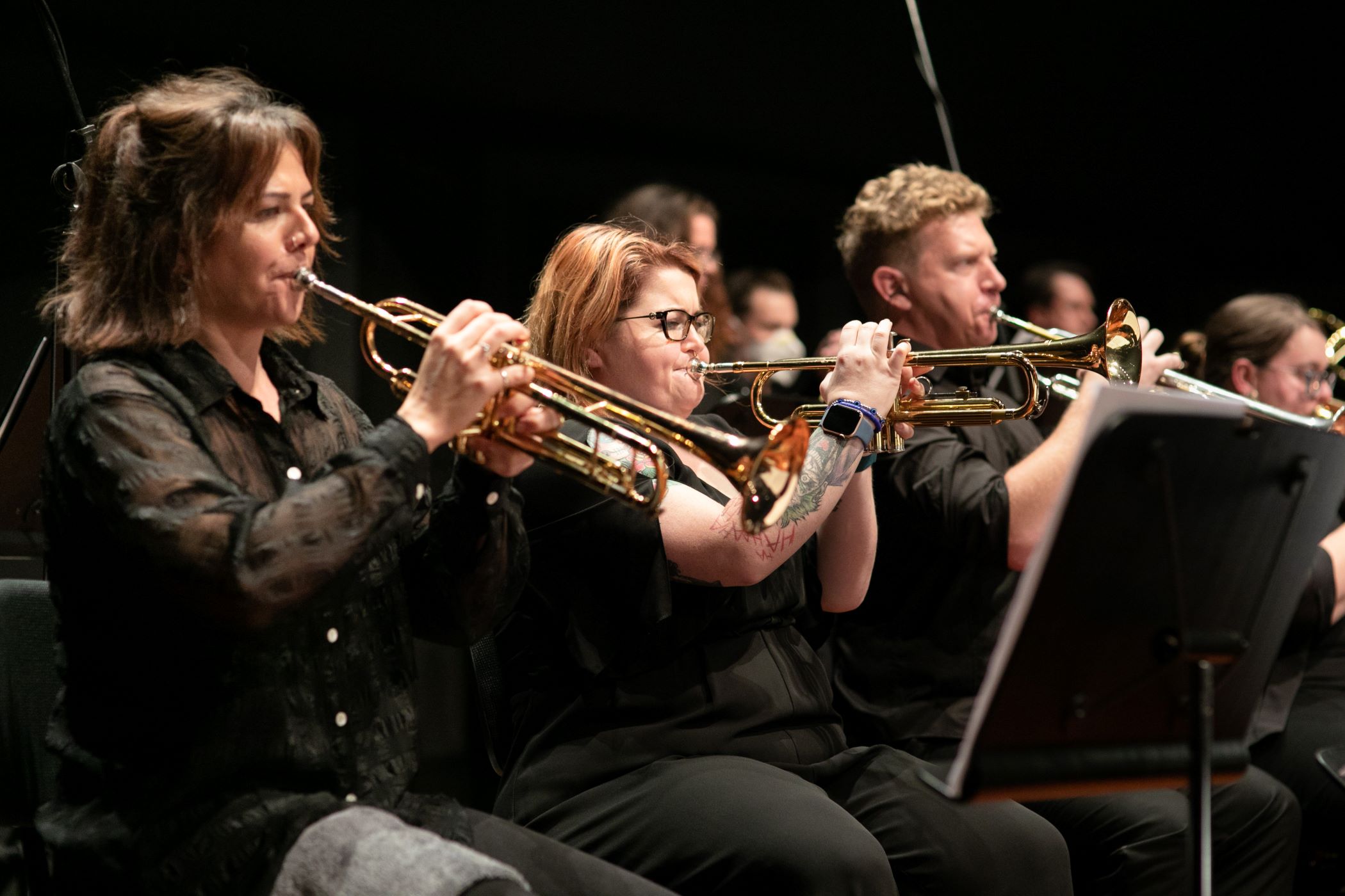 Trumpet players perform as part of an orchestra, seated on stage and reading from music stands.