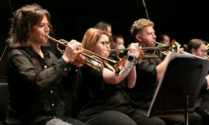 Trumpet players perform as part of an orchestra, seated on stage and reading from music stands.