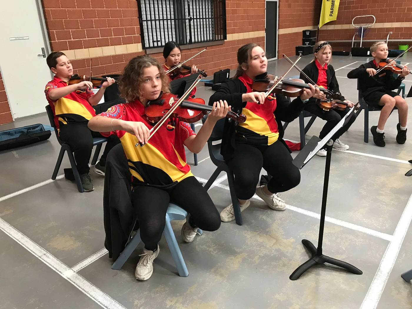 A group of primary school students performing as a string ensemble in a school assembly area.