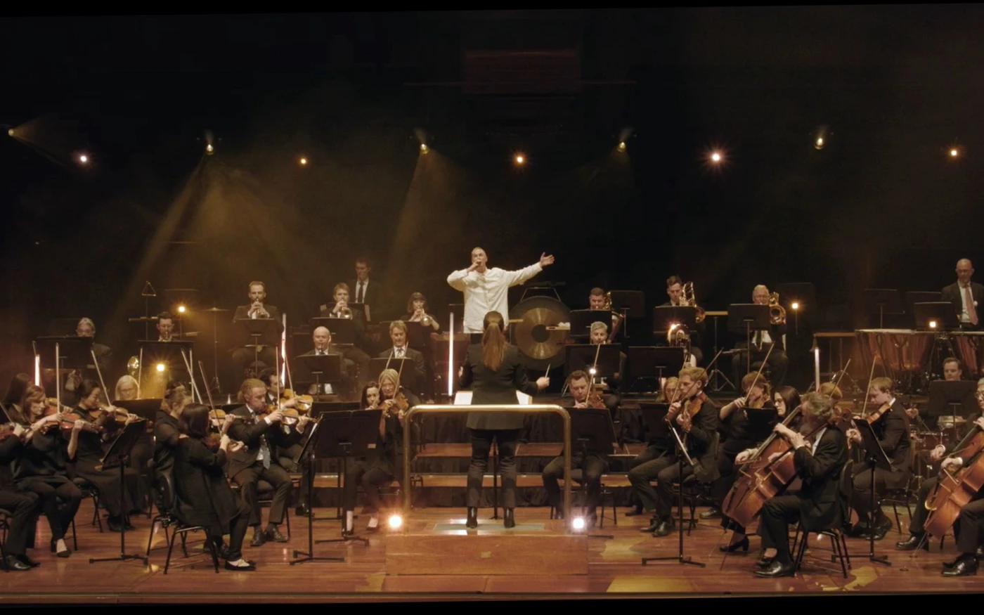 Still from 'Jimmy Recard' music video: Drapht performs on stage with WASO, under dramatic lighting in a dark concert hall.