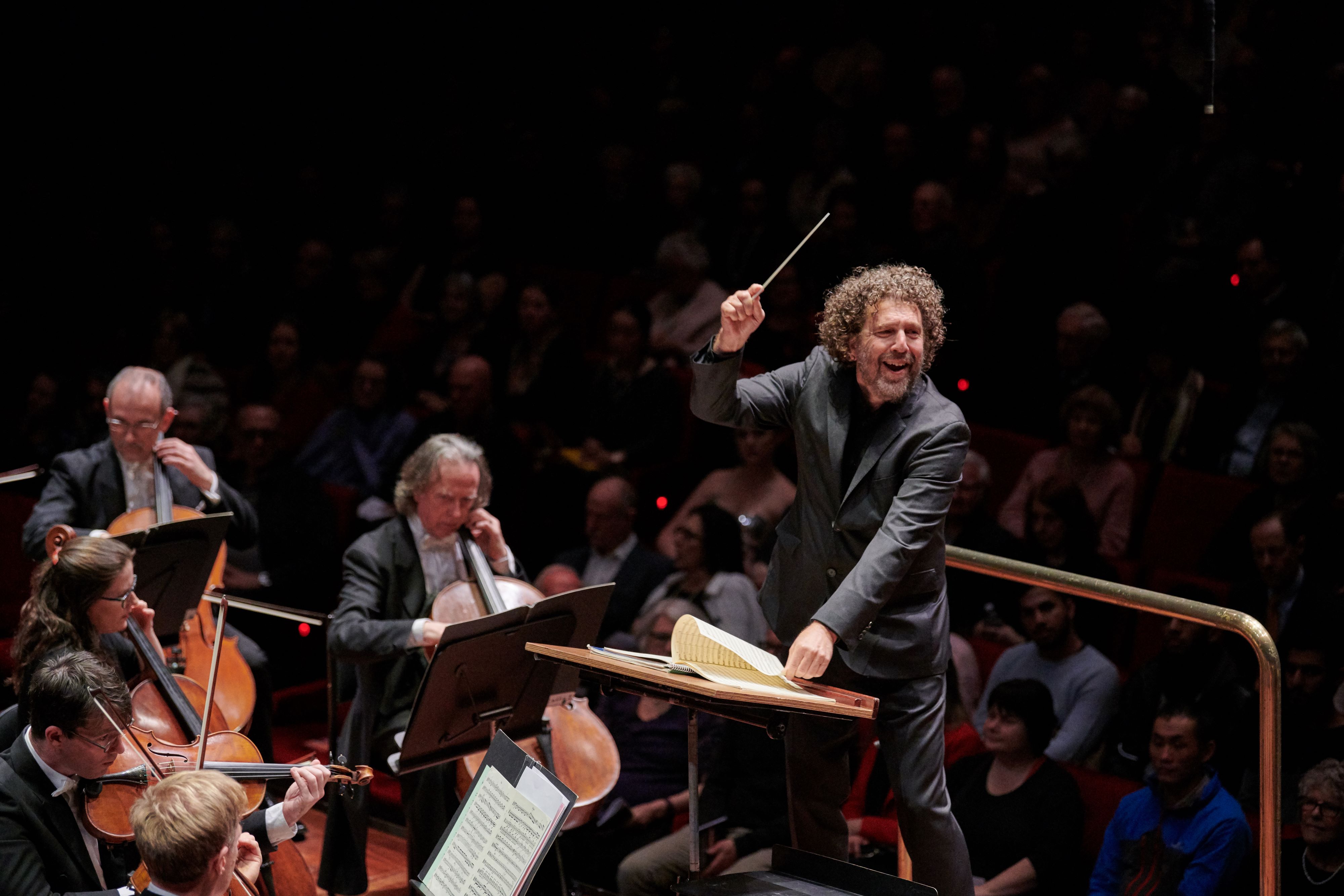 Asher Fisch smiling while conducting an orchestra.