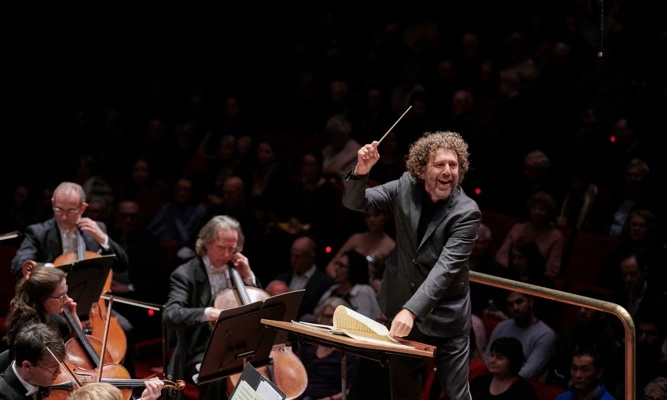 Asher Fisch smiling while conducting an orchestra.