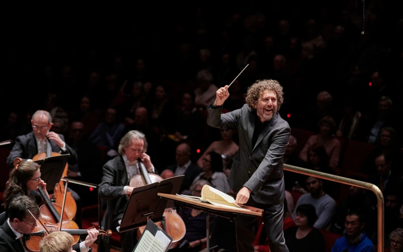 Asher Fisch smiling while conducting an orchestra.