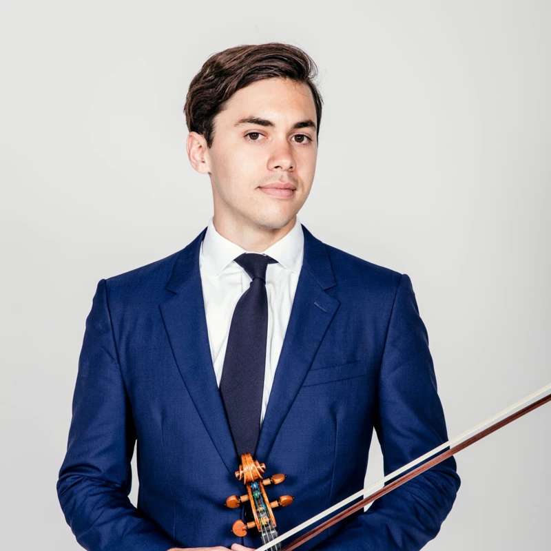 Headshot of Benjamin Beilman wearing a navy suit jacket and tie, holding a violin.