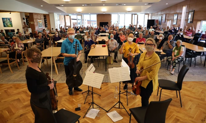 Four WASO string musicians stand before a large, joyous audience in a retirement village hall.